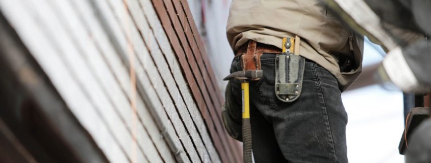 A man with tools stands outside of a home in need of repairs.