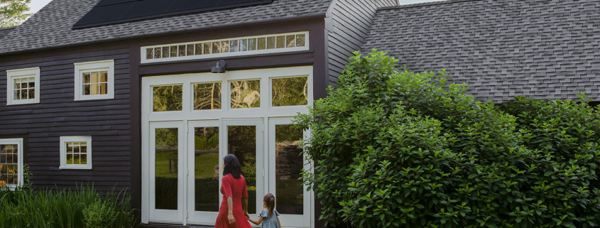 A woman holding the hand of a young girl, walking in front of a dark gray home with solar panels on the rooftop.