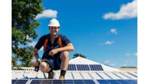 A construction man holding a drill and smiling while crouching on a rooftop with solar panels