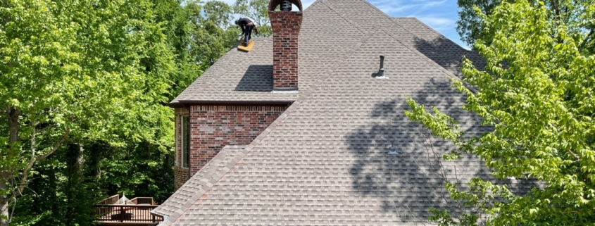 A roofer puts the finishing touches on the roof of a large brick house in spring or summertime. Large trees with bright green leaves stand on either side of the house. The sky beyond is bright blue with a few wispy clouds.