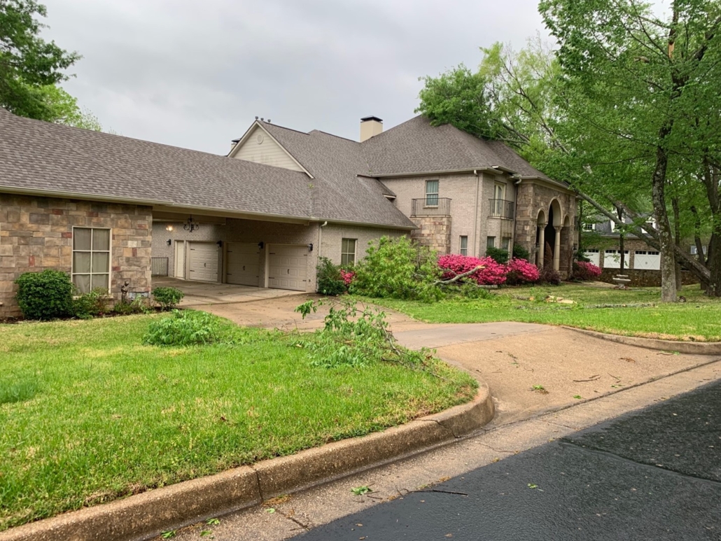 A tree that has fallen on the roof of a large home. Branches from the tree have landed all over the yard, near the garage, and past the carport.