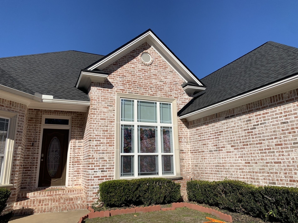 A brick house with dark roof shingles pictured in the daytime. 